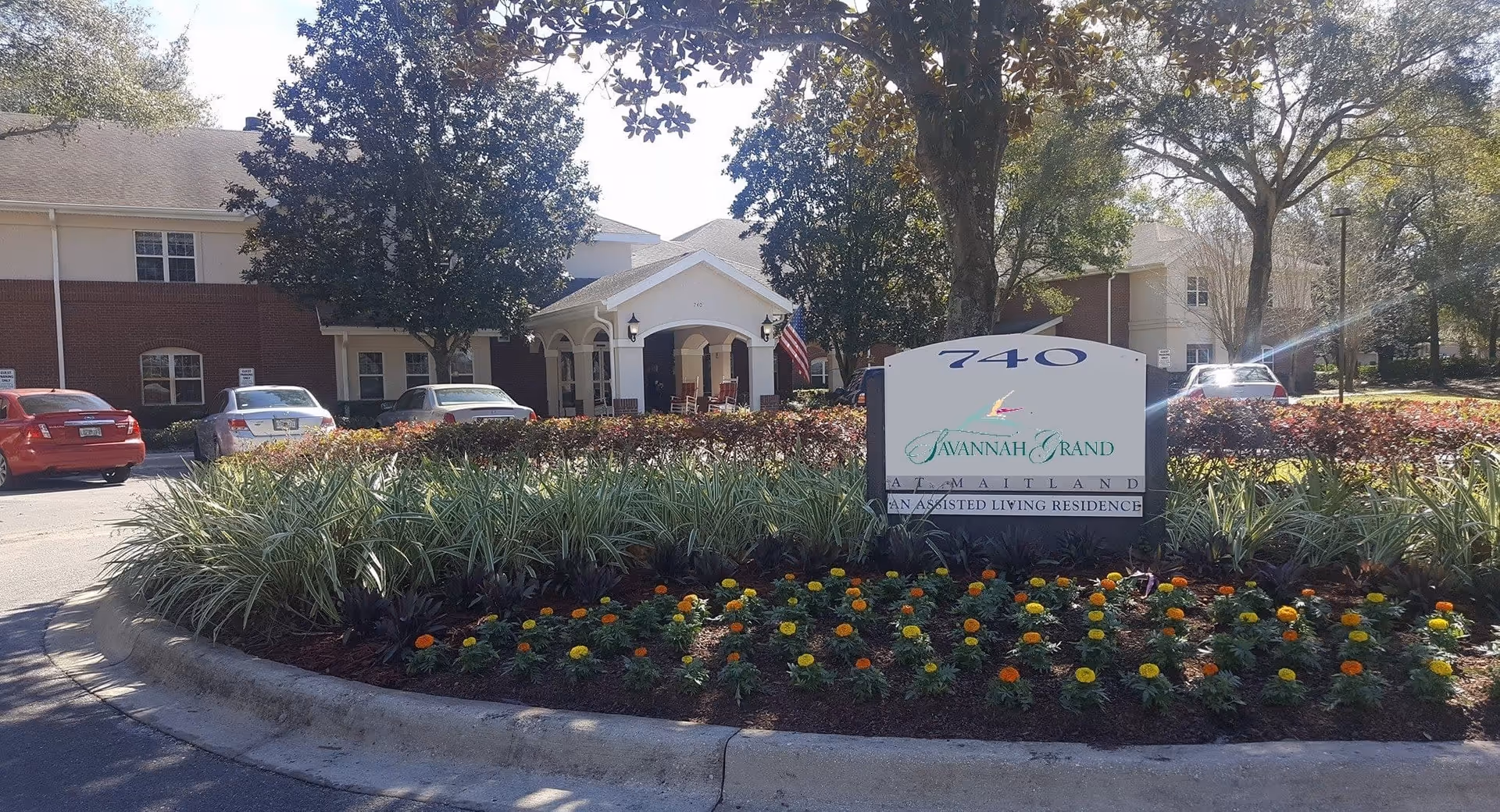 Exterior view of a senior living facility with a landscaped flower bed in front. The flower bed has yellow and orange flowers arranged to spell out 'AMBULANCE'. Behind the flower bed is a sign that reads '740 Savannah Grand at Maitland An Assisted Living Residence'. The building has a brick and white facade with a covered entrance, rocking chairs on the porch, and several parked cars in the parking lot. Trees and shrubs surround the area under a clear sky.