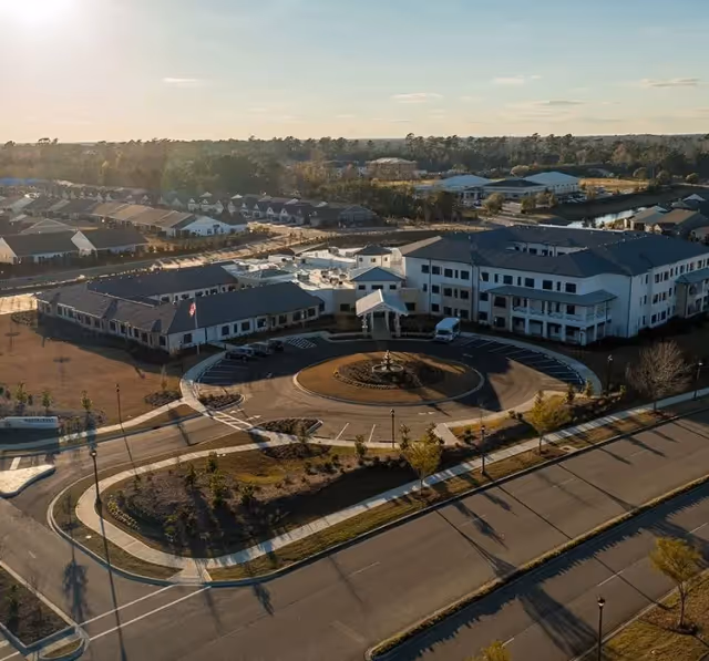 Aerial view of Watercrest Myrtle Beach senior living facility showing a large building complex with a circular driveway and fountain in front, surrounded by landscaped areas and roads, with residential houses and trees in the background under a clear sky.