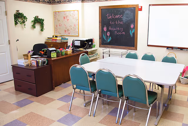 A room with a white rectangular table surrounded by eight green chairs. On the wall, there is a chalkboard with the message 'Welcome to the Activity Room' written in pink chalk with drawings of flowers. There is a wooden desk with office supplies, a microwave, and some books. The floor has a checkered pattern with beige, purple, and brown tiles. The walls are decorated with two green hanging plants and a floral picture.