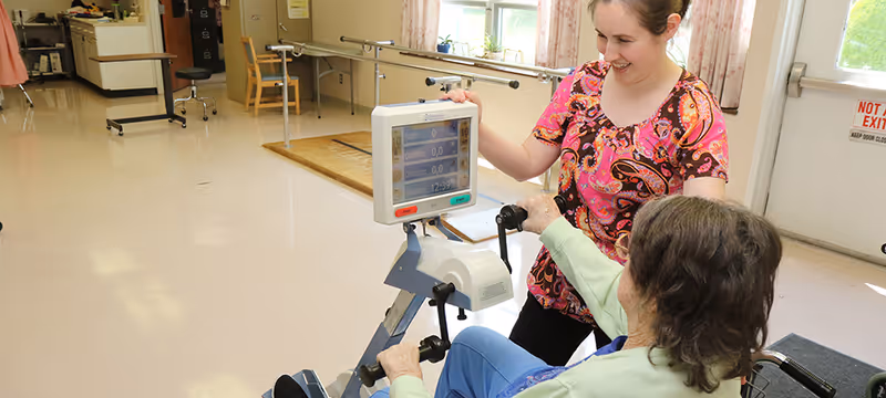 A caregiver in a colorful shirt assists an elderly person in a wheelchair with using a stationary exercise bike in a bright room with handrails along the walls and windows letting in natural light.