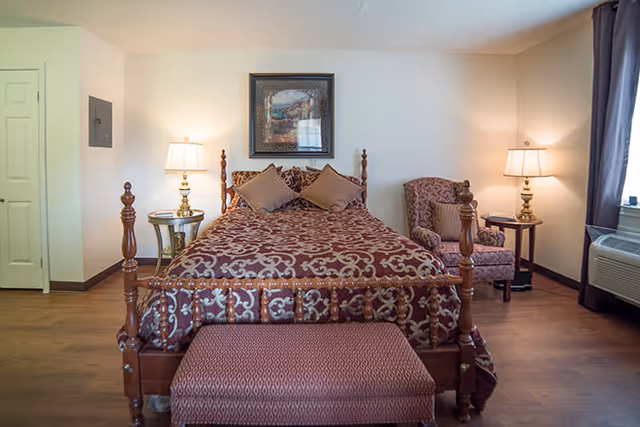 A bedroom with a wooden bed frame featuring a patterned maroon and beige bedspread and two matching pillows. There is a cushioned bench at the foot of the bed, a framed painting on the wall above the bed, two bedside tables each with a lamp, and a patterned armchair with a pillow next to a window with curtains. The room has wooden flooring and light-colored walls.