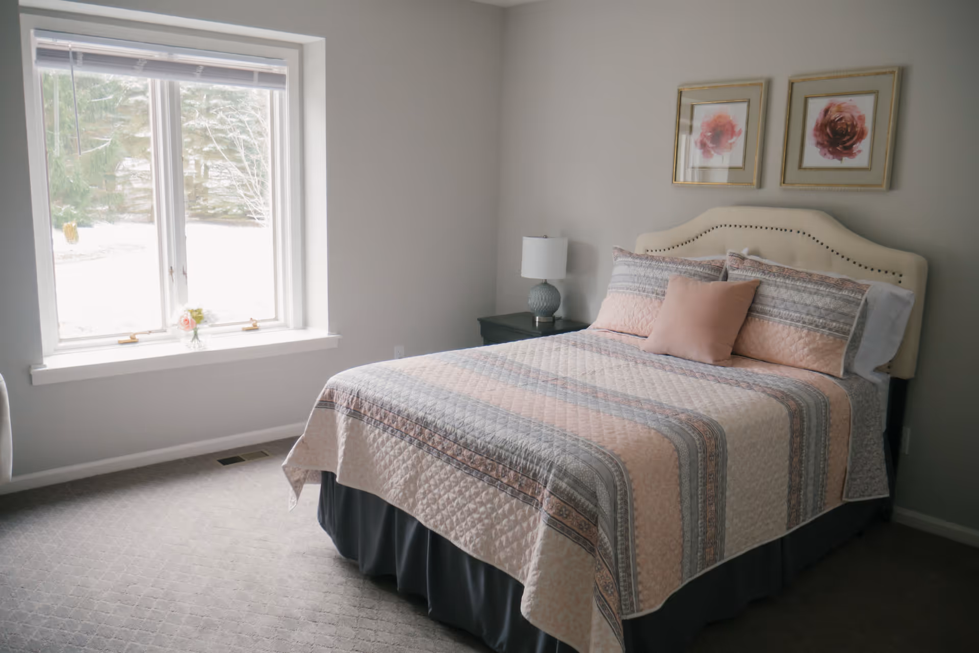 A bedroom with a neatly made bed featuring a patterned quilt in soft pink and gray tones, two framed floral artworks above the headboard, a bedside table with a lamp, and a large window letting in natural light.