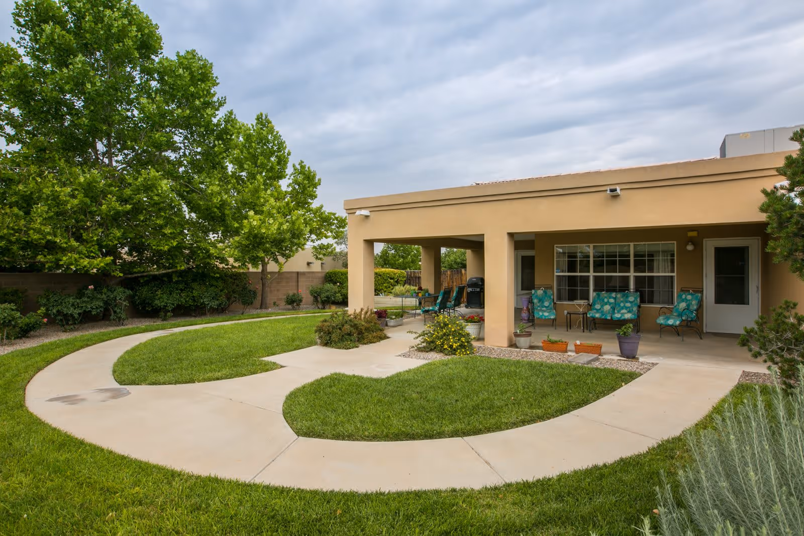 Outdoor patio area with a covered seating space featuring chairs with teal cushions and small tables. The patio is surrounded by a curved concrete walkway and well-maintained green grass, with trees and shrubs along the perimeter under a cloudy sky.
