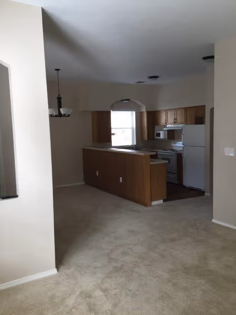 Interior view of a kitchen area in a senior living facility with wooden cabinets, a white refrigerator, stove, microwave, and a countertop with a sink. The kitchen is adjacent to a carpeted dining or living space with a hanging light fixture.