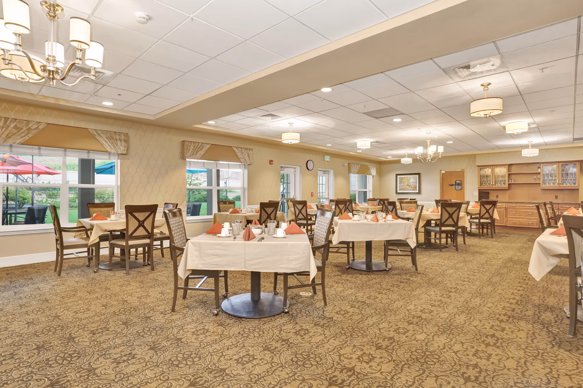 A spacious dining room with multiple tables covered in white tablecloths and set with cups, plates, and folded orange napkins. The room features large windows with beige curtains, allowing natural light to enter. There are wooden chairs around each table, a patterned carpet, and ceiling lights including chandeliers. Outside the windows, colorful umbrellas and greenery are visible.
