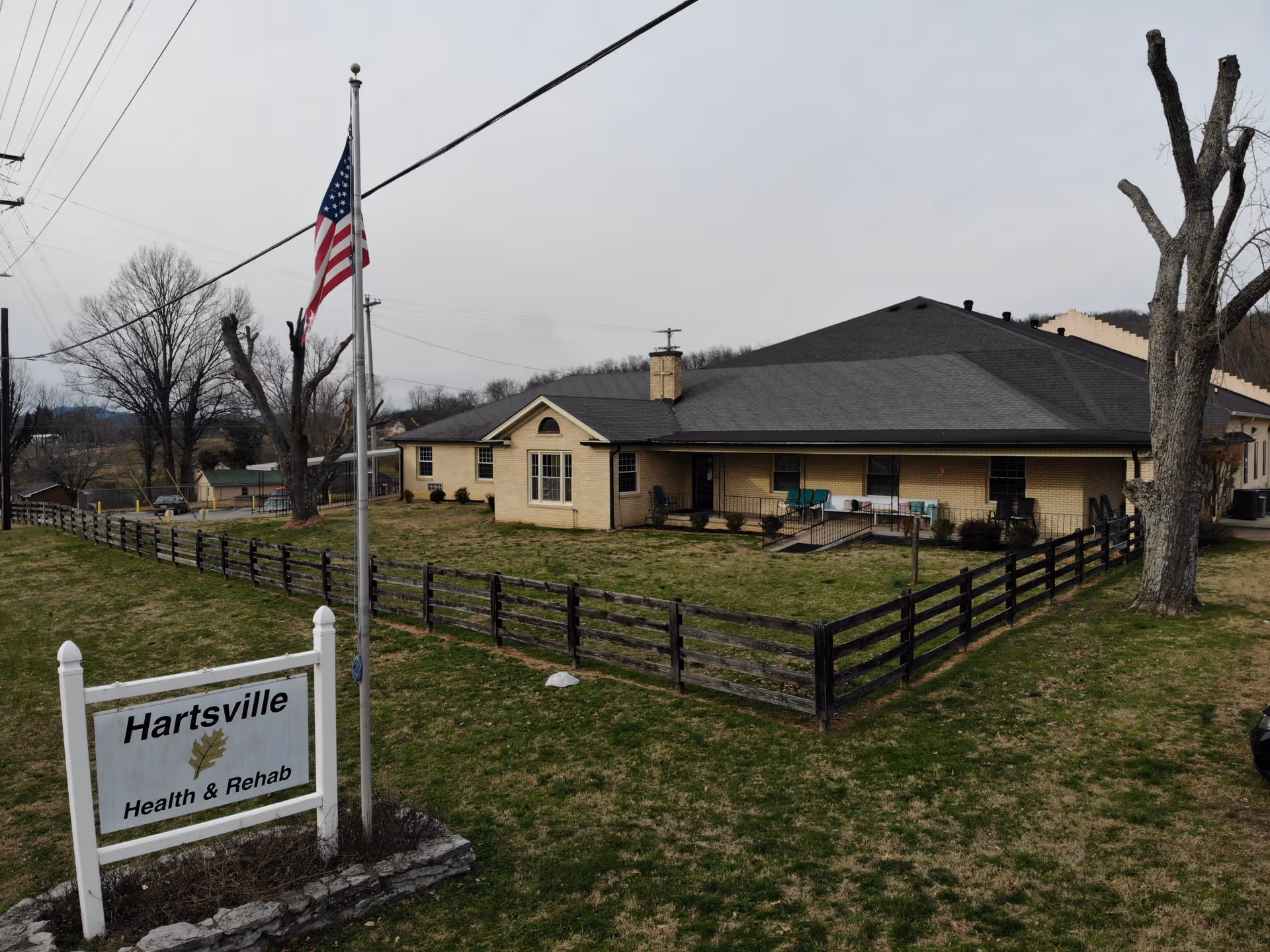 Exterior view of Hartsville Health & Rehab building with a fenced grassy yard, an American flag on a flagpole, and a sign displaying the facility's name. The building is a single-story structure with a dark roof and light-colored walls, surrounded by leafless trees and power lines under a cloudy sky.