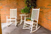 Two white wooden rocking chairs on a concrete floor next to a small white metal table with a potted plant, set against a brick wall corner.