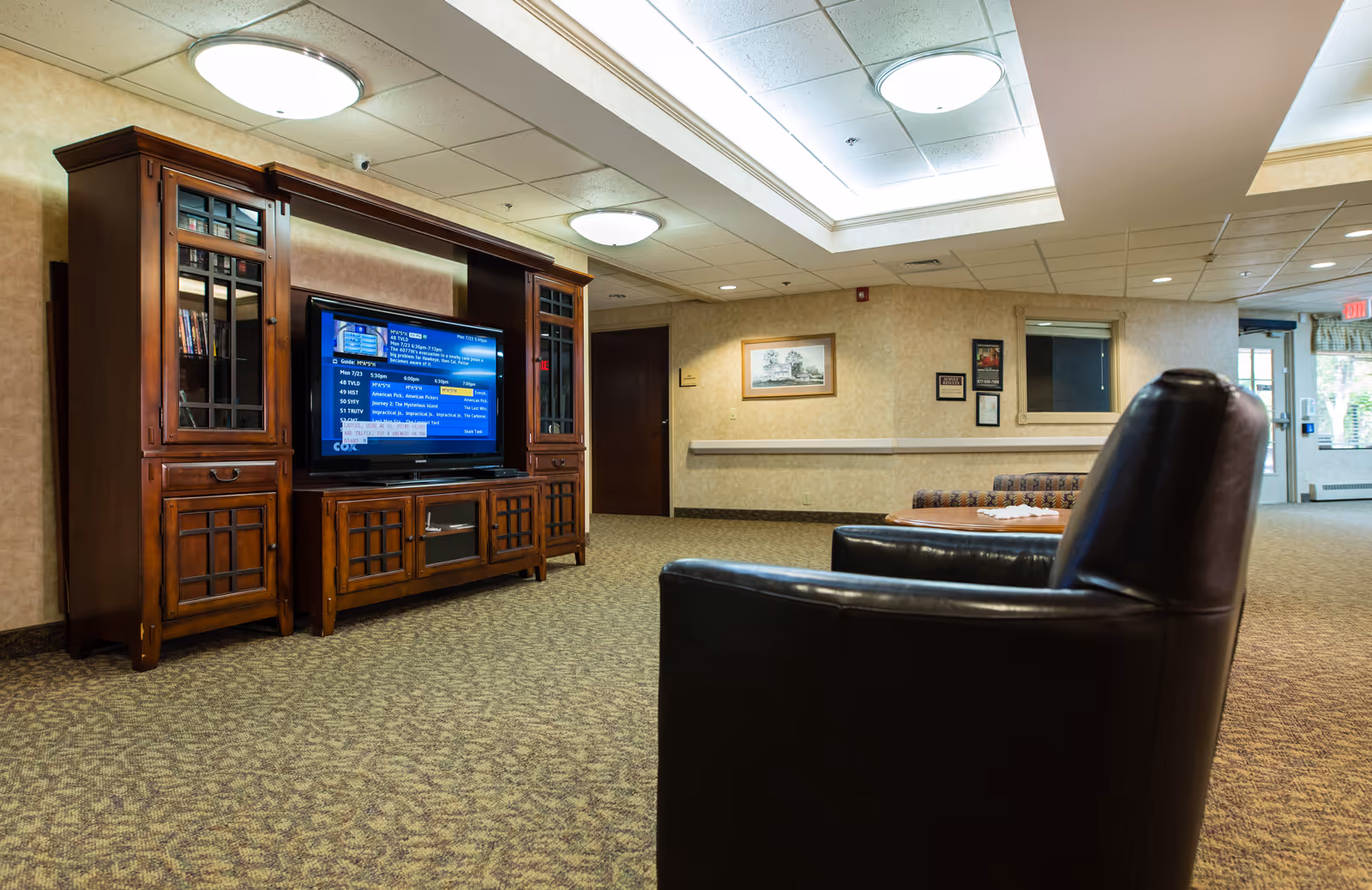 A cozy common area in an assisted living facility featuring a large wooden entertainment center with a flat-screen TV, a black leather armchair in the foreground, patterned carpet, and beige walls with framed artwork. The ceiling has recessed lighting and a skylight, and there is a door and window visible in the background.