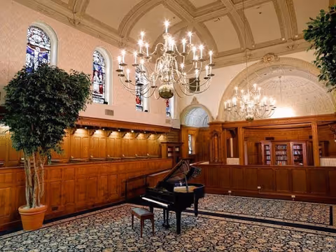 Ornately wood-paneled interior room with a grand piano, chandeliers, stained glass windows and patterned carpet.