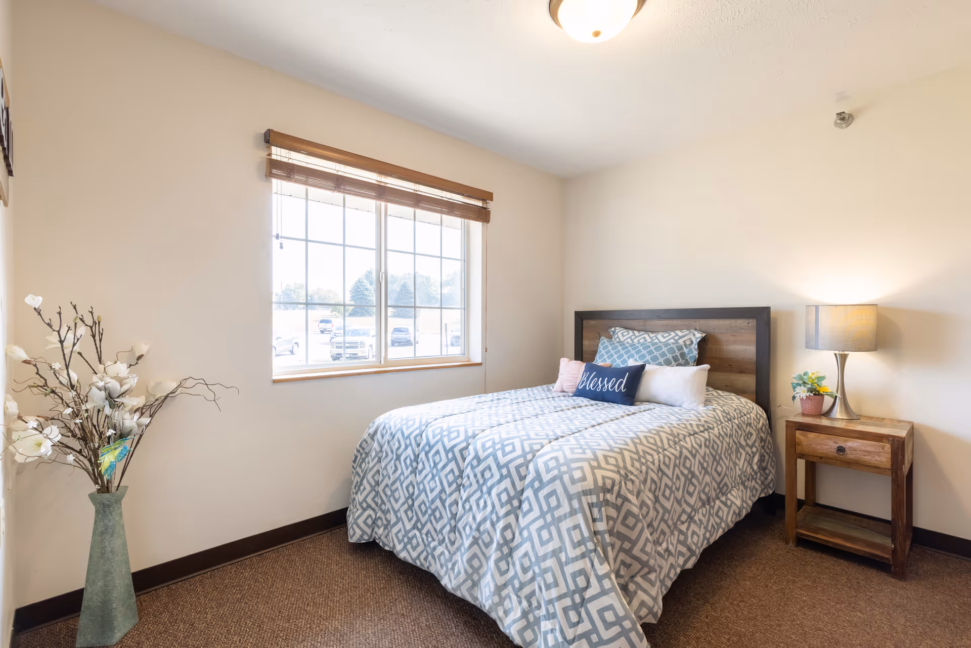 Bright, simply furnished bedroom with a double bed dressed in patterned bedding, a wooden nightstand with lamp, a window, and a decorative floor vase.