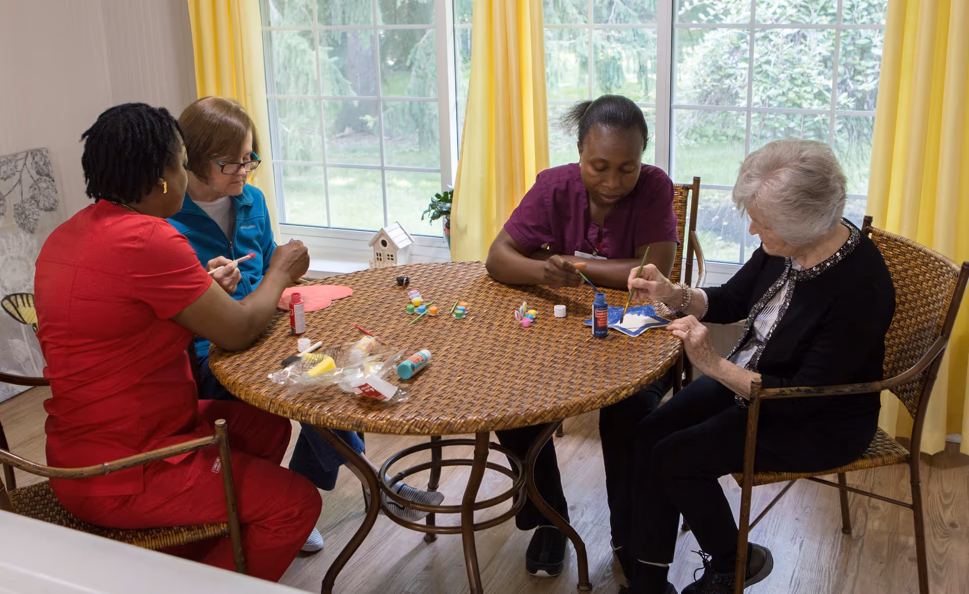 Four women sitting around a round wicker table engaged in arts and crafts activities, painting and using glue. The room has large windows with yellow curtains and a view of greenery outside.