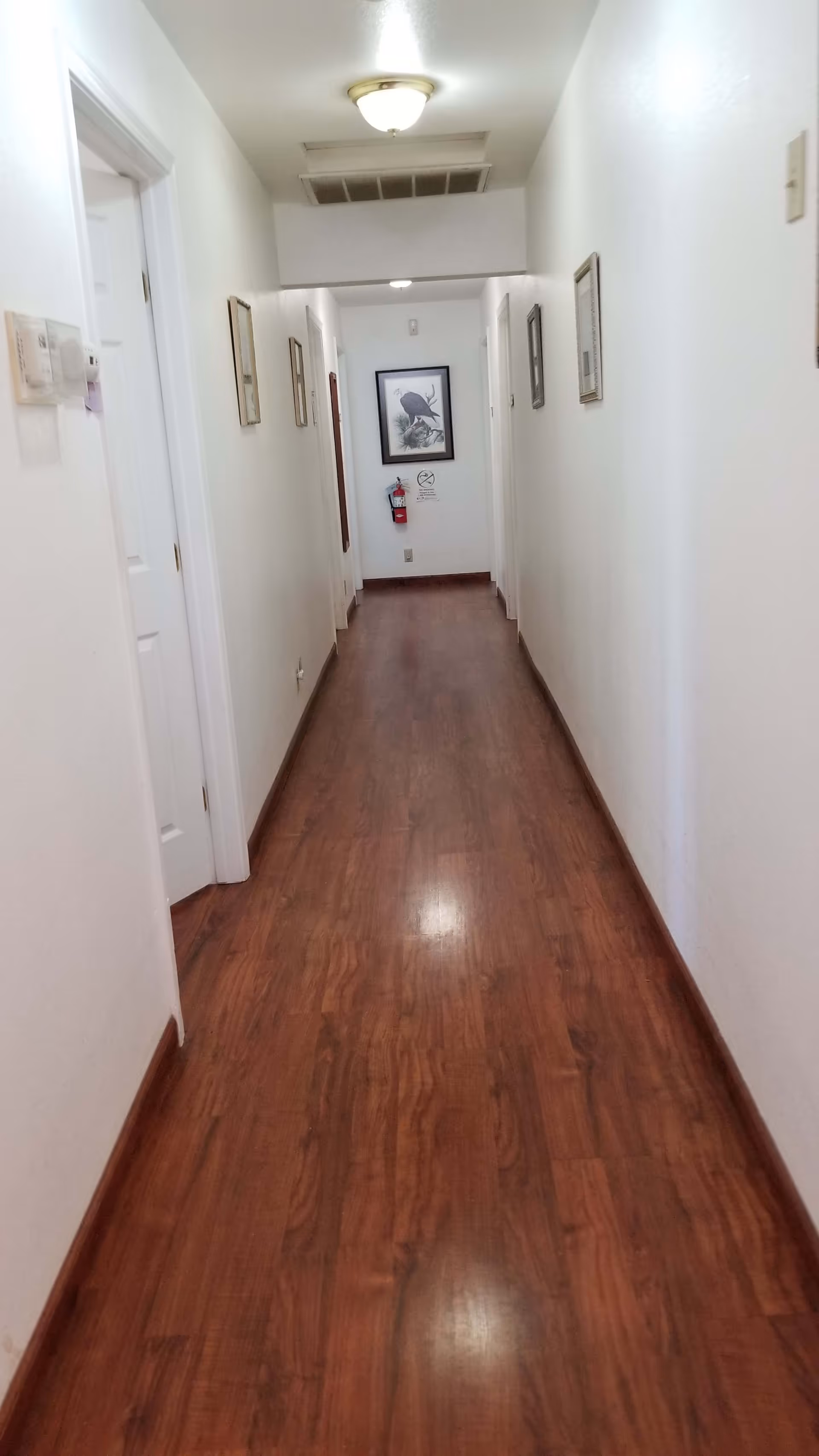 A long, narrow hallway with wooden flooring and white walls. Several doors line both sides of the hallway. There are framed pictures hanging on the walls and a fire extinguisher mounted at the end of the hallway beneath a framed picture of a bird. The ceiling has a light fixture and an air vent.