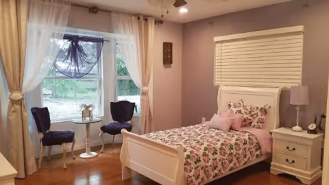 A cozy bedroom with lavender walls featuring a white single bed with floral bedding and pink pillows. Next to the bed is a white nightstand with a lamp and clock. Two blue upholstered chairs and a small round table are positioned near a window with sheer white curtains and a decorative purple swag. The room has wooden flooring and a ceiling fan.