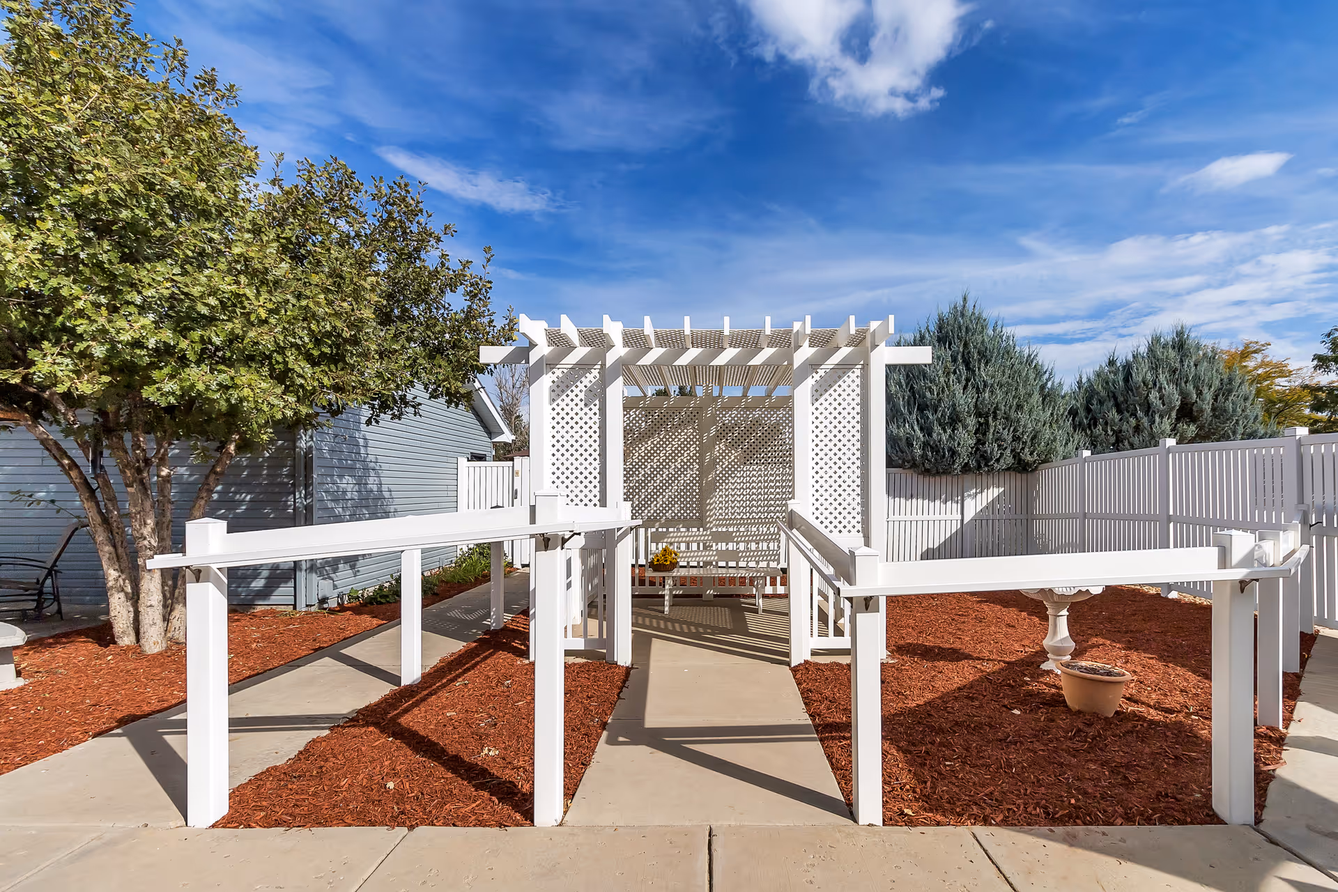 Outdoor garden area with a white wooden pergola and lattice panels, surrounded by mulch beds and a concrete pathway with white handrails. Trees and shrubs are visible in the background under a blue sky with some clouds.
