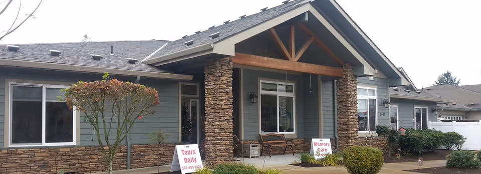 Exterior view of a single-story senior living facility with stone and wood accents, a covered entrance supported by stone pillars, and signs indicating daily tours and memory care services.