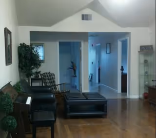 Interior view of a living room area with wooden flooring, a piano with a bench, a black leather chair, a rocking chair, a small table, and a plant. The room opens into a hallway with doors and framed pictures on the walls.