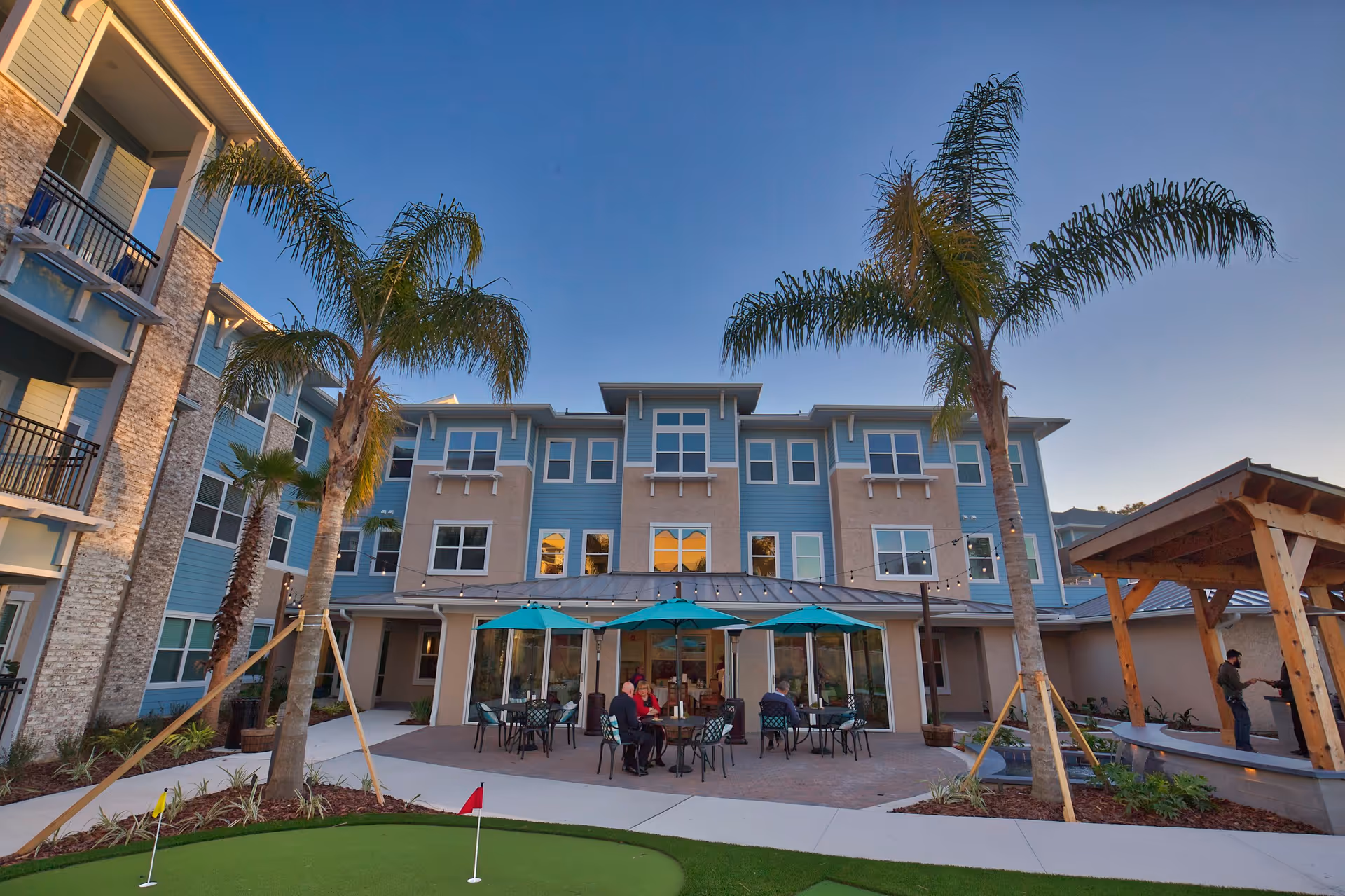 Outdoor courtyard area of a senior living facility with a putting green in the foreground, several palm trees, and a three-story building in the background. There are tables with teal umbrellas where people are seated, and a wooden pergola structure on the right side.