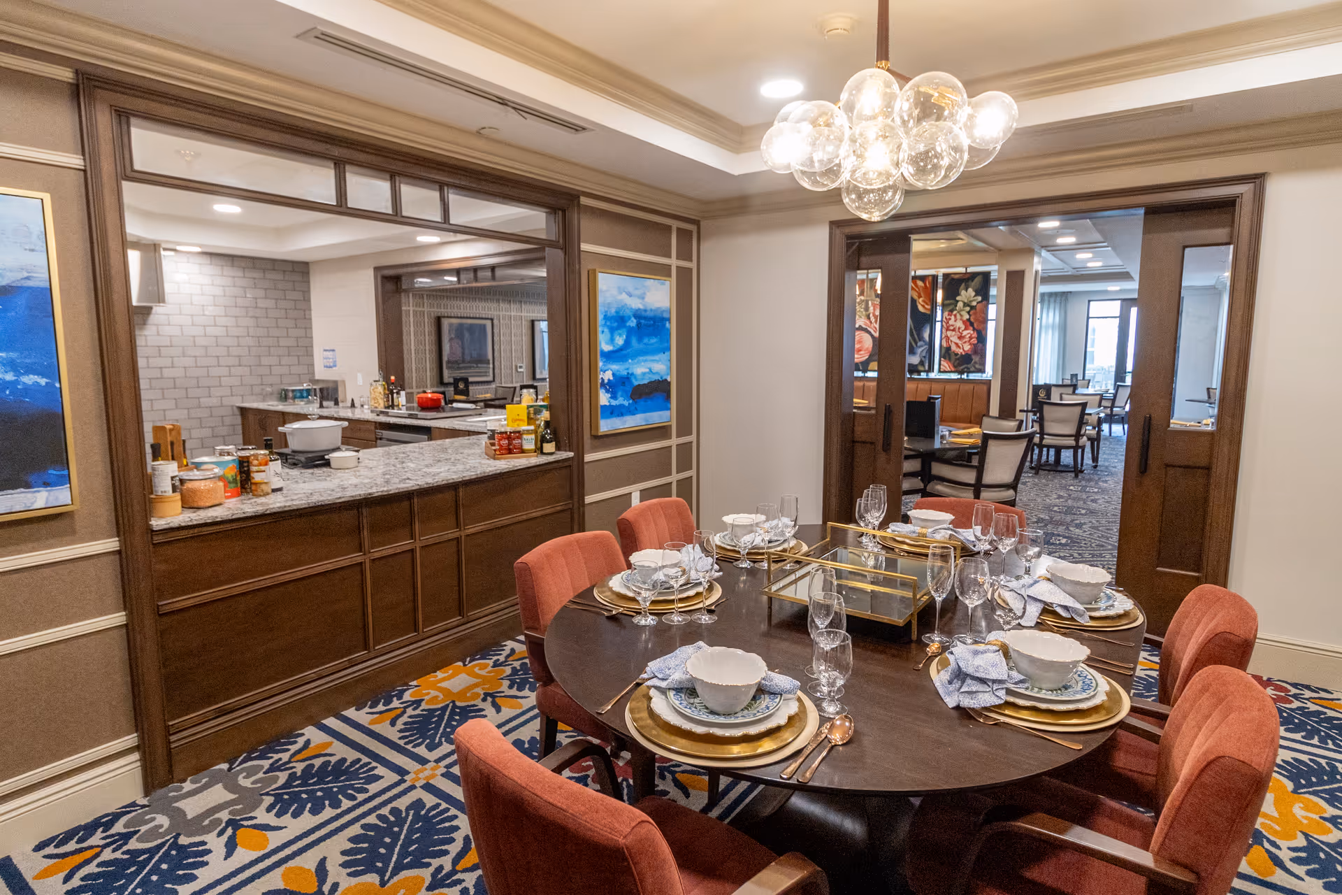 A furnished dining room with a round table set for six, red upholstered chairs, a chandelier, and a pass-through window to a kitchen area.