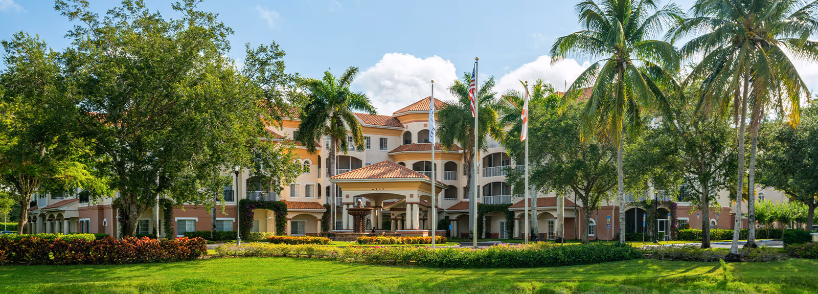 Front exterior view of Terracina Senior Living facility with a large entrance canopy, multiple flags, palm trees, and well-maintained landscaping including green grass and shrubs under a partly cloudy sky.
