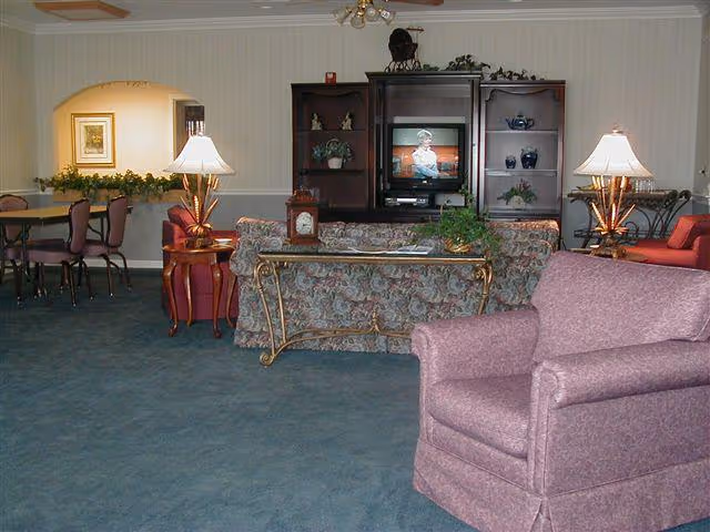 A cozy living room area in a senior living facility featuring a floral patterned sofa, a purple upholstered armchair, two side tables with lamps, a wooden entertainment center with a TV, and a dining area with a table and chairs in the background.