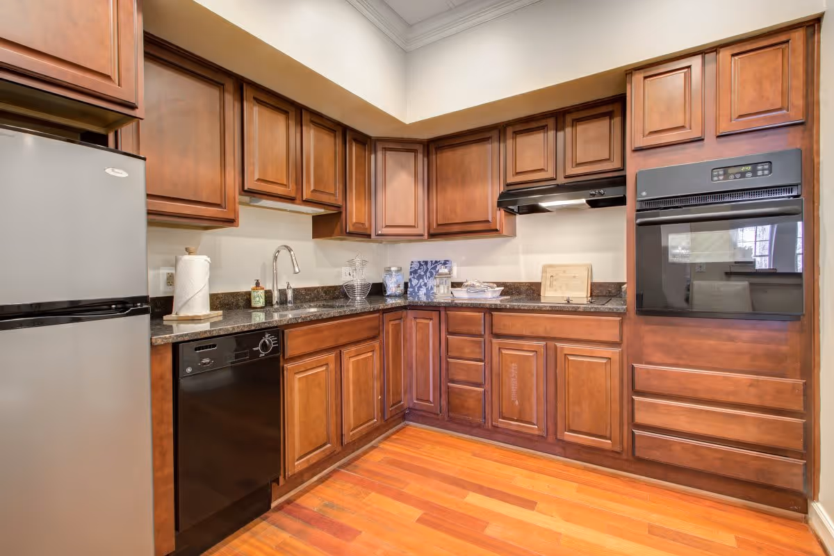 A kitchen with wooden cabinets, a stainless steel refrigerator, a black dishwasher, a built-in oven, a sink with a faucet, and granite countertops. The floor is made of polished wood.