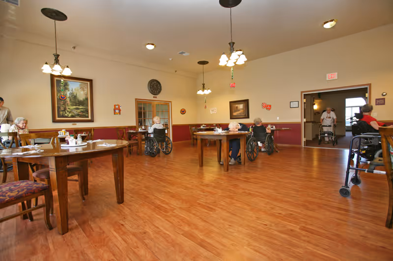 A spacious dining room in a senior living facility with wooden floors and several round wooden tables. Elderly residents, some in wheelchairs, are seated at the tables or moving around. The walls are decorated with framed paintings and a clock. Ceiling lights hang from above, and an exit sign is visible above a doorway in the background.