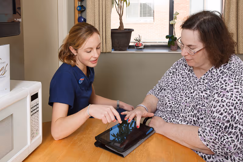 A caregiver and an older woman sit at a table indoors as the caregiver shows her something on a tablet.