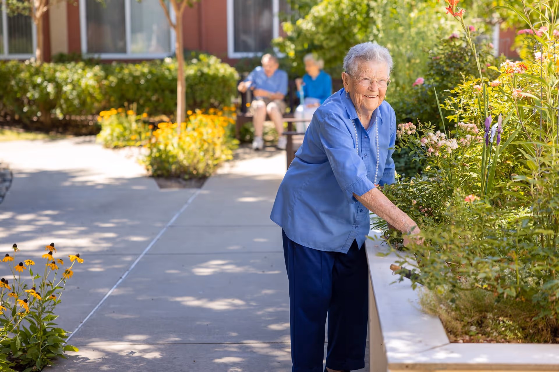 An elderly woman wearing a blue shirt and dark pants is tending to plants in a garden area outside a senior living facility. In the background, two other elderly individuals are seated on a bench near a building with windows, surrounded by greenery and flowers.