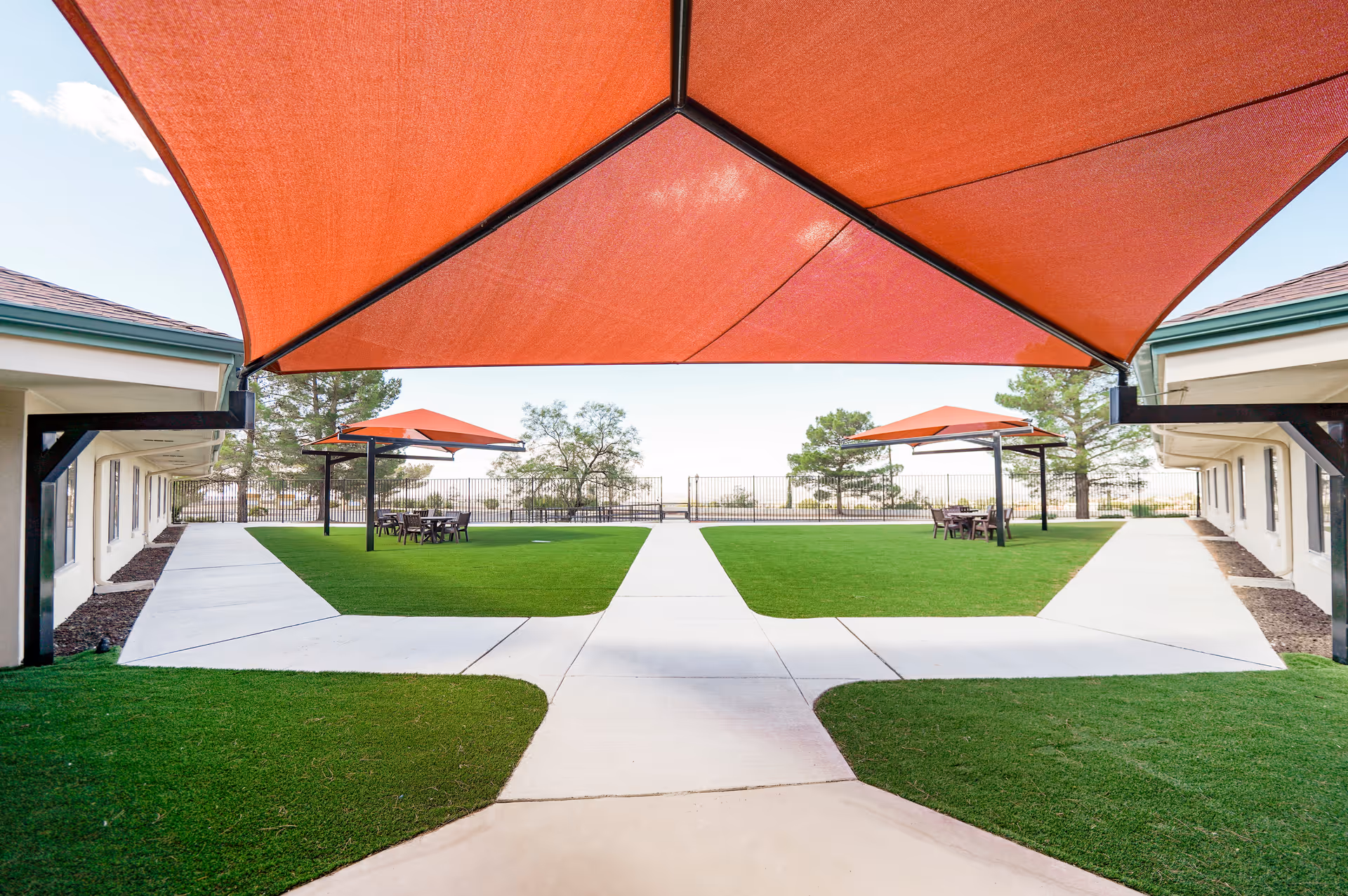 A landscaped courtyard with intersecting concrete walkways, green lawns, red shade sails and outdoor seating between two single-story buildings.