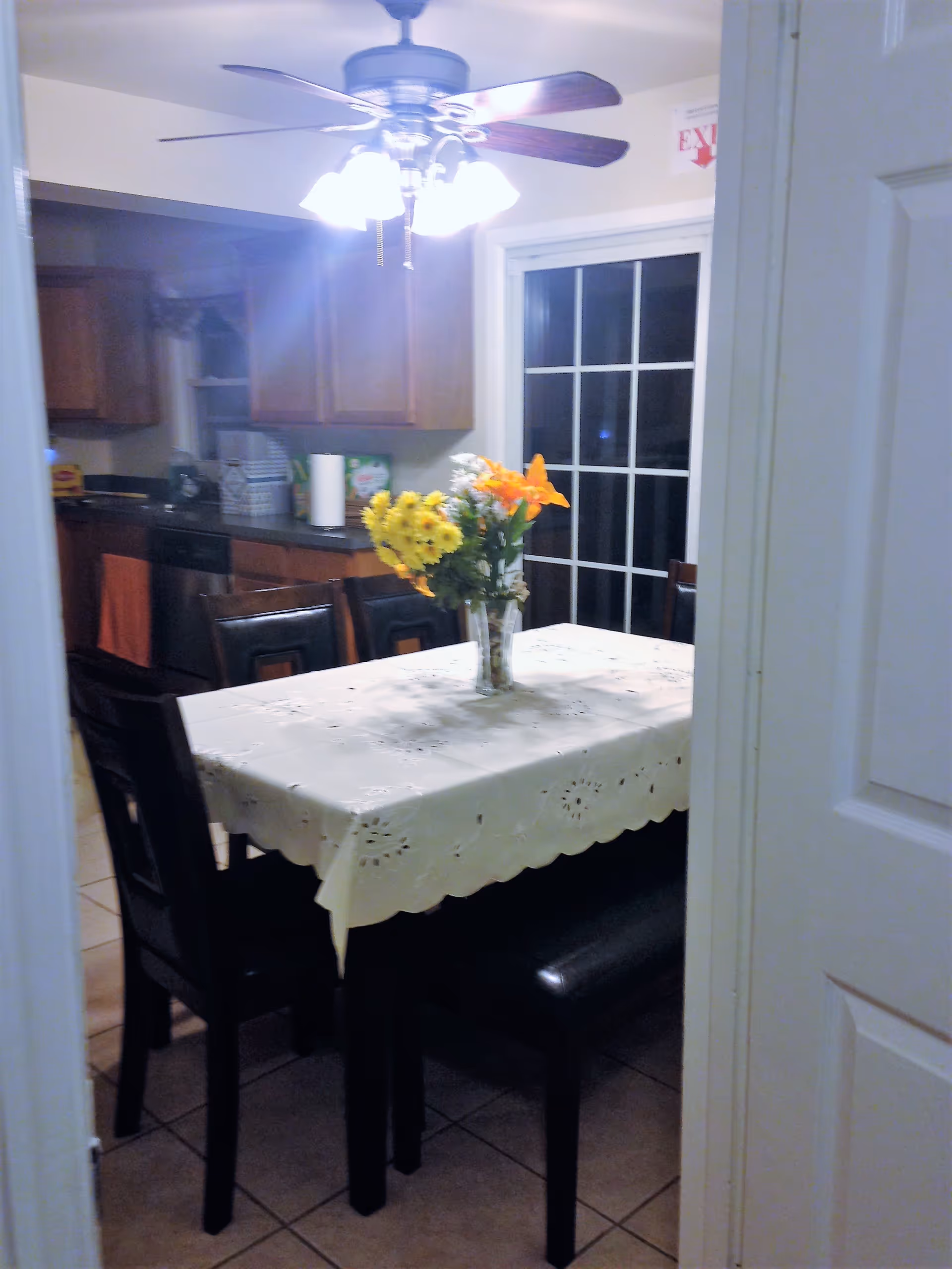 View through a doorway into a dining area with a rectangular table covered with a white tablecloth and a vase of yellow and orange flowers on top. The table is surrounded by dark chairs and a bench. In the background, there is a kitchen with wooden cabinets, a countertop with various items, and a ceiling fan with lights above the table. A sliding glass door is visible behind the table.
