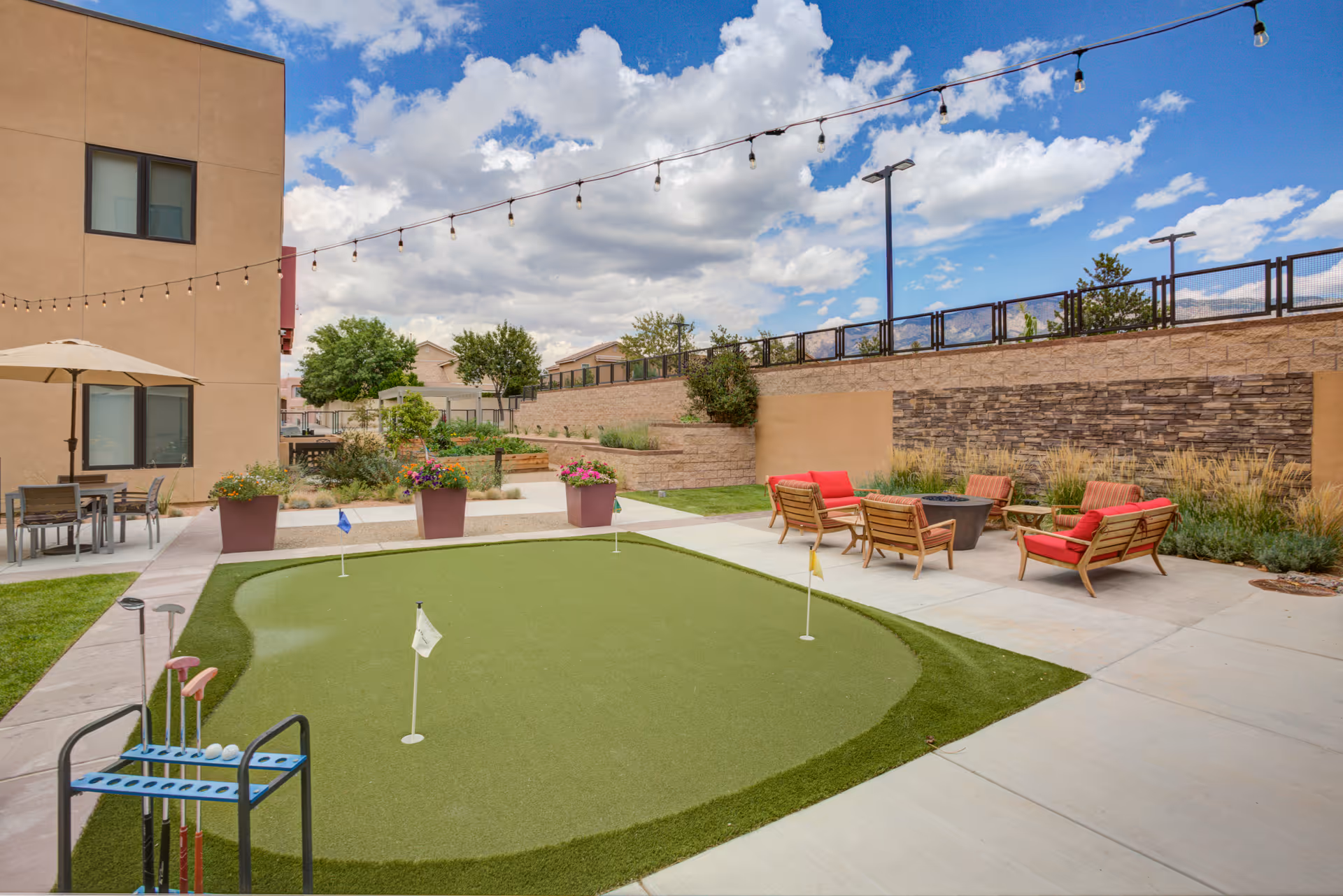 Outdoor patio area at Amaran Senior Living featuring a small artificial putting green with golf clubs and balls, surrounded by concrete walkways. There are several wooden chairs with red cushions arranged around a fire pit. String lights are hung above the area, and there are large potted plants and a beige building wall in the background under a partly cloudy blue sky.