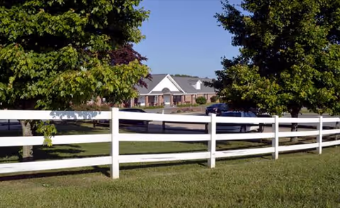 White wooden fence frames a grassy yard with trees and a facility building in distance.