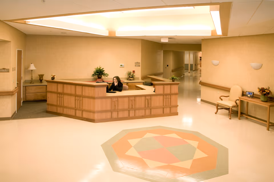 A reception desk in a senior living facility lobby with a seated receptionist and a patterned floor medallion.