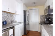Bright galley kitchen with white cabinets, stainless steel refrigerator and appliances, gray speckled countertops and a mosaic tile backsplash.