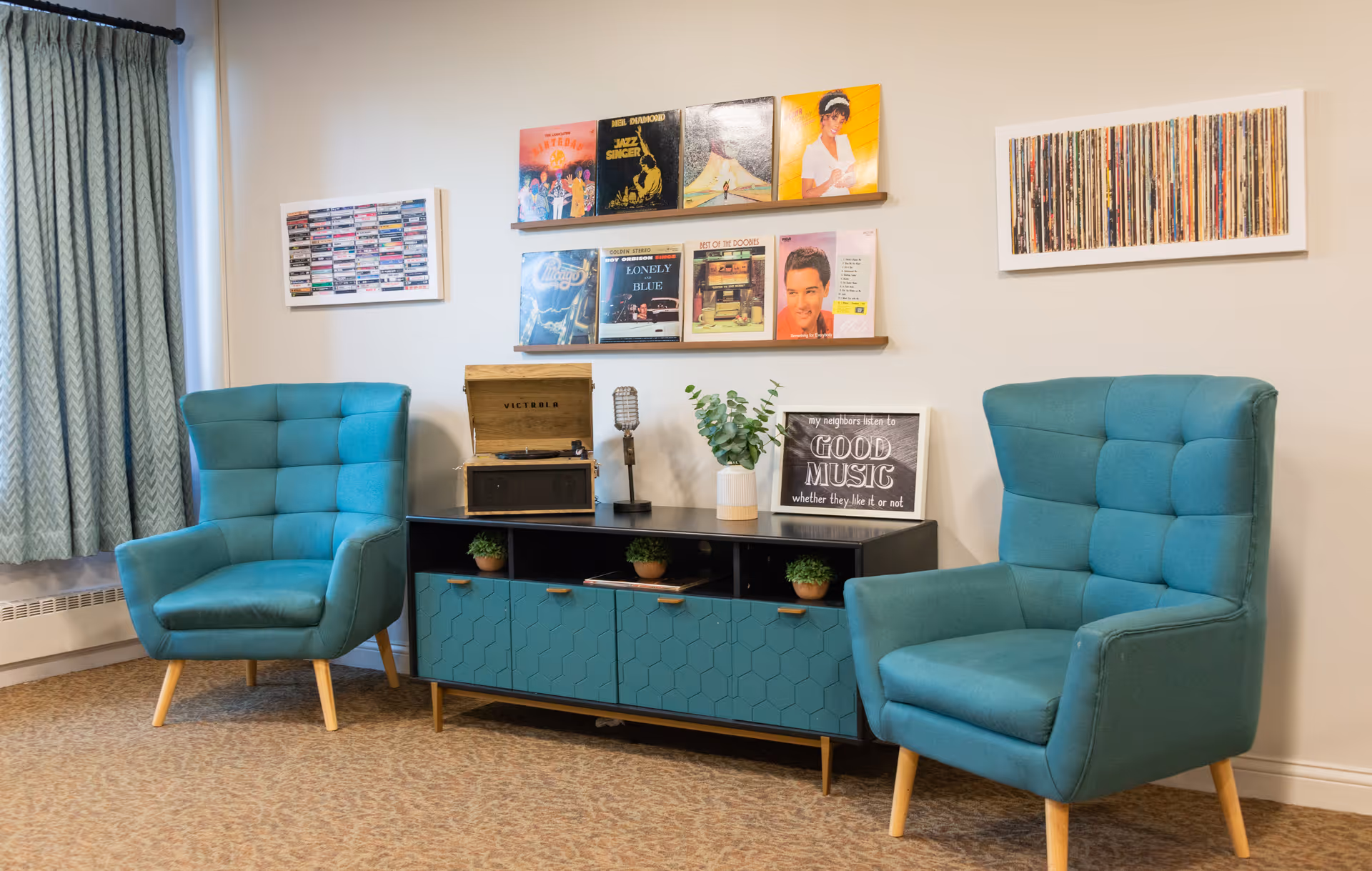 A cozy sitting area with two teal upholstered armchairs facing each other. Between them is a teal and black cabinet with small potted plants on top. On the cabinet sits a vintage-style record player, a decorative microphone, a small plant, and a framed sign that reads 'my neighbors listen to GOOD MUSIC whether they like it or not.' Above the cabinet are two wooden shelves displaying vinyl record covers. The walls have framed artwork featuring cassette tapes and vinyl records. A window with light blue curtains is visible on the left side.