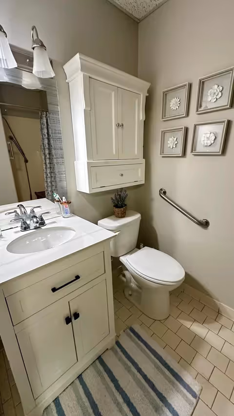 Small bathroom with a white vanity and sink, toilet under a wall cabinet, grab bar on the wall, decorative framed art, and a striped floor rug.