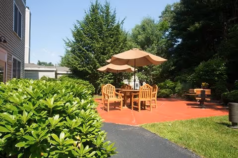 Outdoor patio area with a red paved surface featuring a wooden table and chairs under two large beige umbrellas. The patio is surrounded by green bushes, grass, and tall trees, with part of a building visible on the left side under a clear blue sky.
