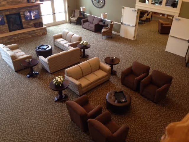 A spacious assisted living facility common area with multiple seating arrangements including beige sofas, brown armchairs, round wooden tables with floral centerpieces, and a fireplace with a stone surround. The room has large windows letting in natural light and a carpeted floor with a leaf pattern.