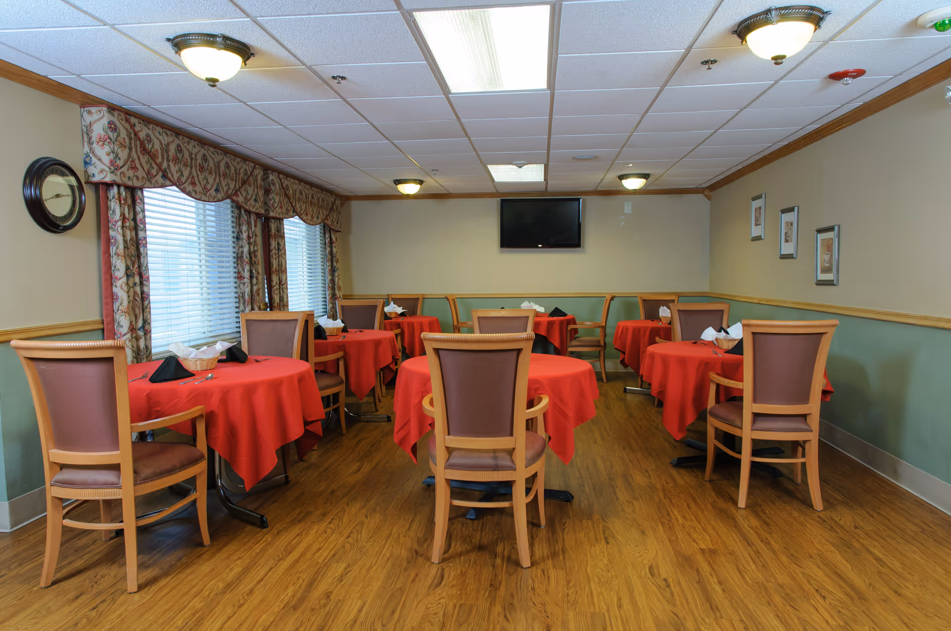 A dining room with several round tables covered with red tablecloths, each set with napkins and baskets. Wooden chairs with cushioned backs surround the tables. The room has wood flooring, beige and green walls, a wall-mounted TV, framed pictures, and windows with floral curtains.