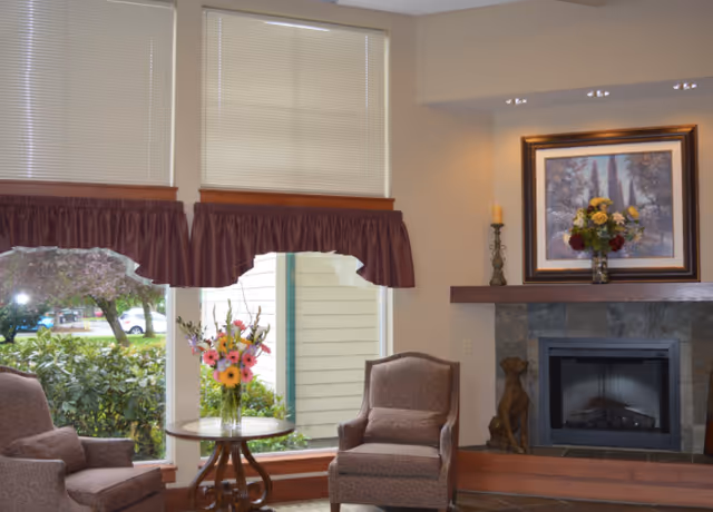 A cozy living room area with two upholstered armchairs facing each other, a round wooden table with a vase of colorful flowers between them, large windows with blinds and maroon valances, and a stone fireplace with a wooden mantle displaying a framed painting and decorative items.