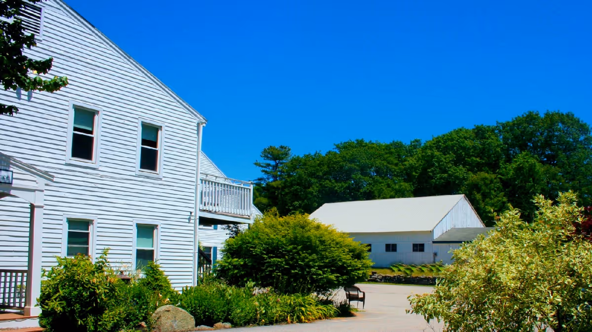 Exterior view of a white two-story building with several windows and a small balcony, surrounded by green bushes and trees under a clear blue sky. Another white building with a slanted roof is visible in the background.