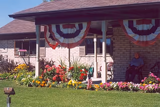 A covered porch area of a brick building decorated with red, white, and blue bunting. There are colorful flowers planted along the edge of the porch, and a person sitting in a wheelchair near the wall. The lawn in front is green and well-maintained.