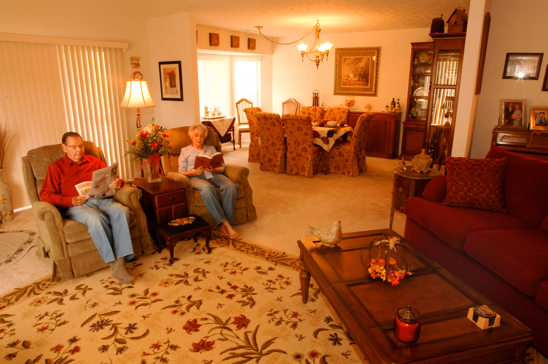 An elderly man and woman sit in armchairs in a warm, well-decorated living room with a coffee table, patterned rug, and dining area visible in the background.