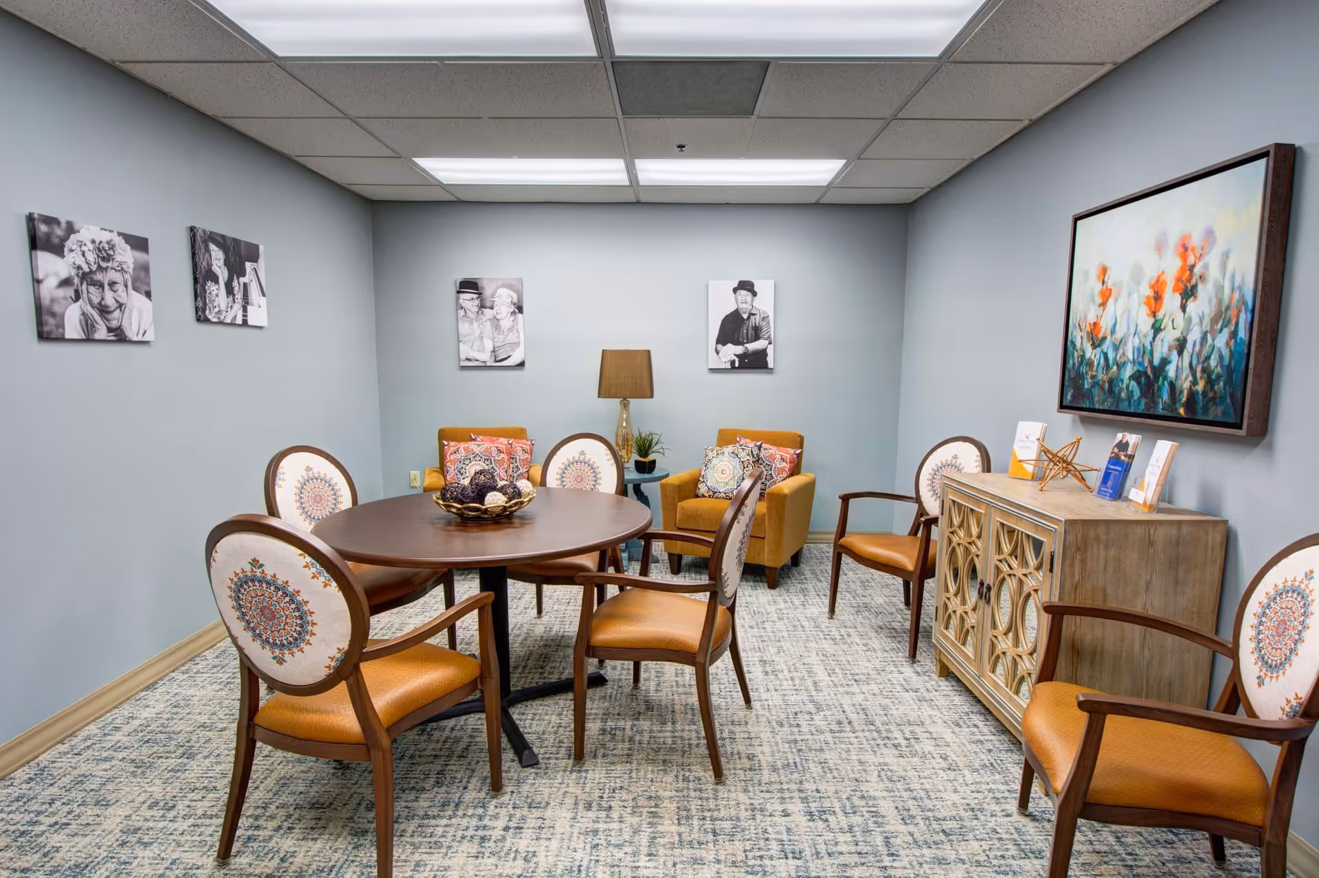 A cozy interior room with a round wooden table surrounded by six chairs with patterned backs and orange cushions. Two orange armchairs with decorative pillows are placed against the far wall, with a lamp and small plant between them. The walls are light gray and decorated with black and white photos of elderly people and a colorful floral painting above a wooden cabinet. The floor is carpeted with a patterned design.