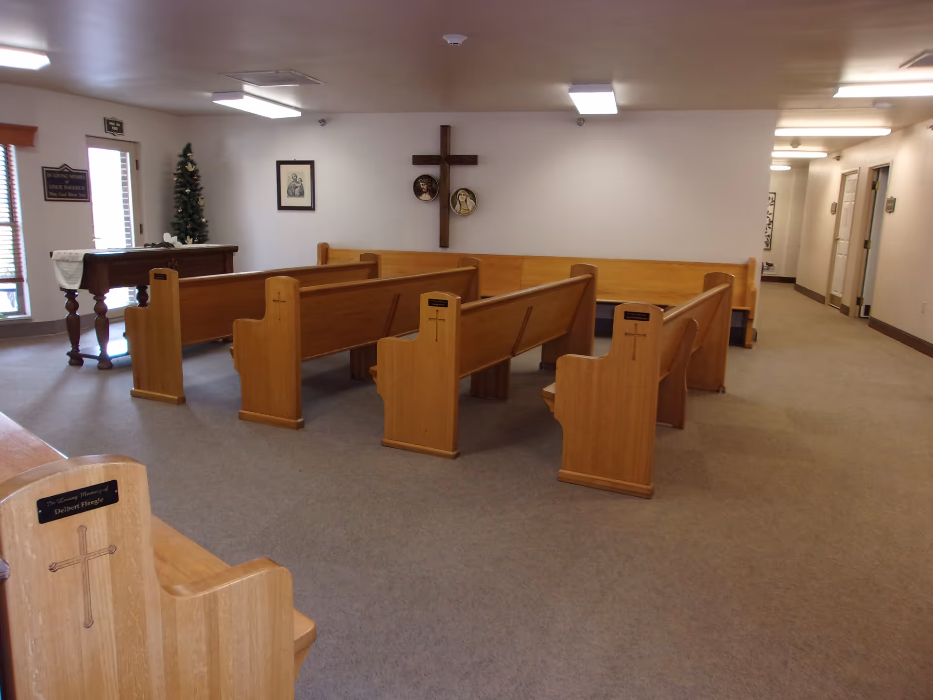 Interior room with wooden pews arranged facing a wall with a large wooden cross and religious artwork. There is a small table with a cloth and a small decorated Christmas tree near the windows. The room has carpeted floors and fluorescent ceiling lights.