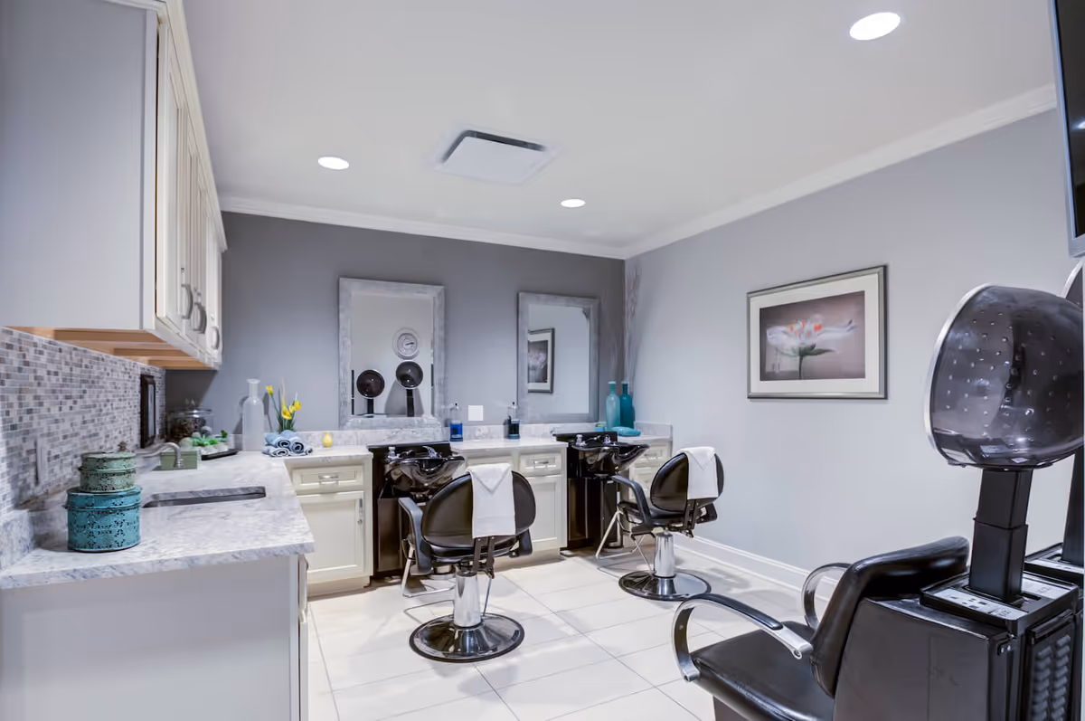 Interior of a salon area in Arbor Terrace Morris Plains featuring two black salon chairs with white towels draped over the backs, two wash basins with mirrors above them, a hair dryer chair, and a countertop with decorative items and cabinets. The walls are painted gray and there is a framed flower picture on one wall.