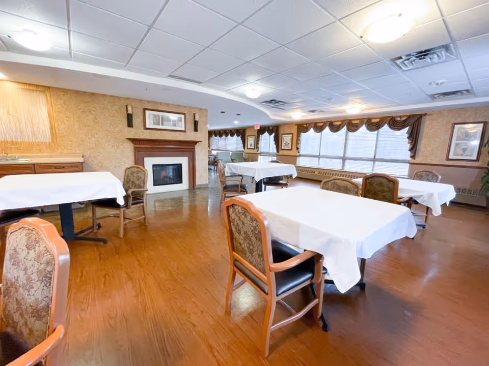 A dining room with several tables covered in white tablecloths and wooden chairs with patterned upholstery. The room has large windows with brown valances, a wooden floor, and a fireplace with a wooden mantle. The walls are decorated with framed pictures and there is a ceiling with recessed lighting.