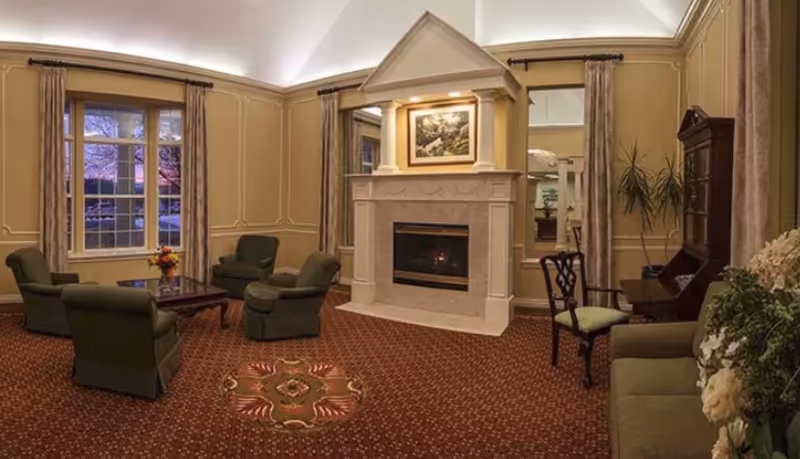 A cozy living room with four green armchairs arranged around a wooden coffee table, a fireplace with a decorative mantle and a painting above it, beige walls with molding, patterned carpet, large windows with curtains, a wooden desk with a chair, and some indoor plants.