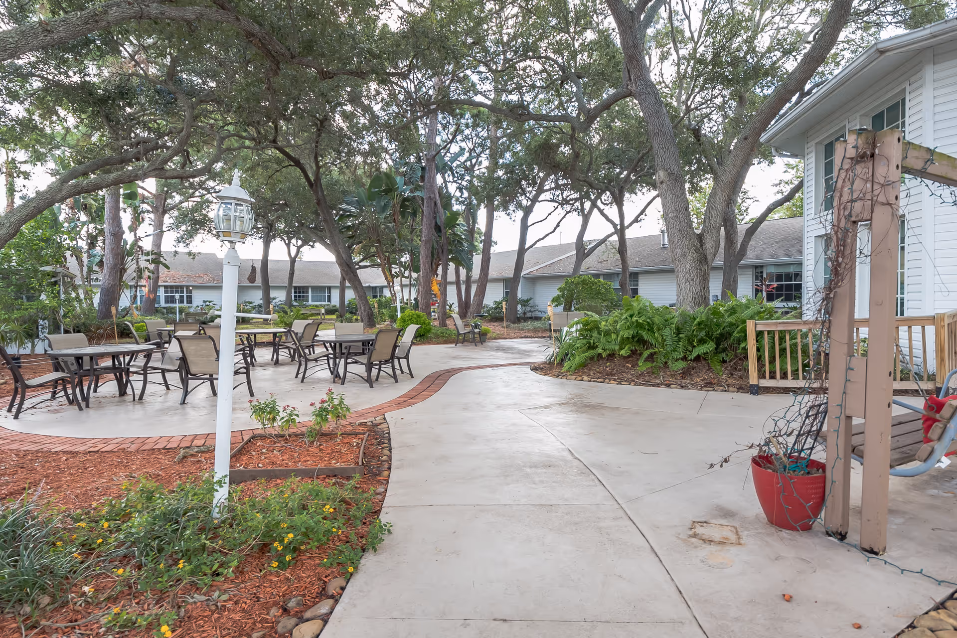 Outdoor patio area at The Pointe with several tables and chairs arranged on a concrete surface surrounded by trees, plants, and landscaped garden beds. A white lamp post and a wooden swing with string lights are also visible.
