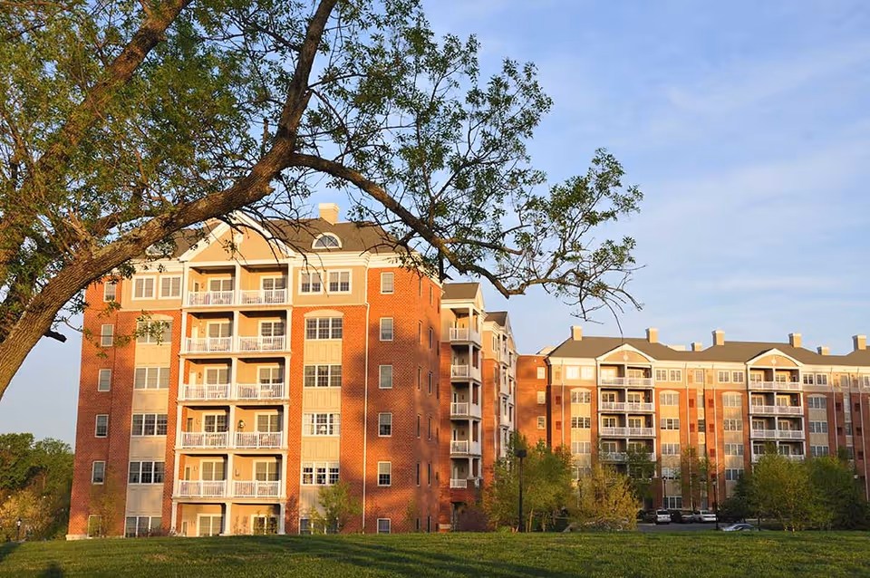 Exterior view of a multi-story senior living facility building with red brick and beige siding, balconies, and large windows, surrounded by green grass and trees under a clear blue sky.